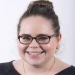 A white woman smiles at the camera. She has glasses and brown hair in a bun.