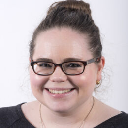 A white woman smiles at the camera. She has glasses and brown hair in a bun.