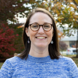 Woman with shoulder-length brown hair shown from shoulders up, wearing blue sweater and brown glasses.