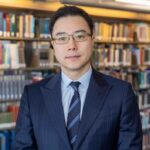 Professor Nathaniel Taeho Yu takes a photo in front of bookshelves at the library with a smile