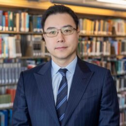 Professor Nathaniel Taeho Yu takes a photo in front of bookshelves at the library with a smile