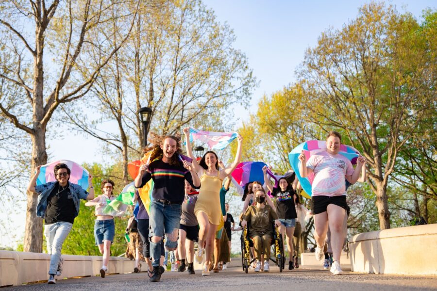 people on the JMU campus running toward the camera, smiling, and holding various pride flags.