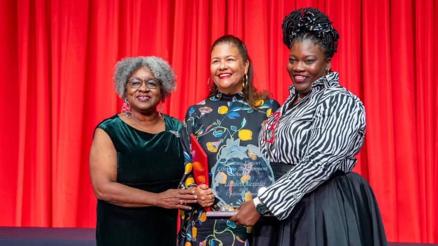 Three women, one holding an inscribed glass award, stand in front of a red curtain